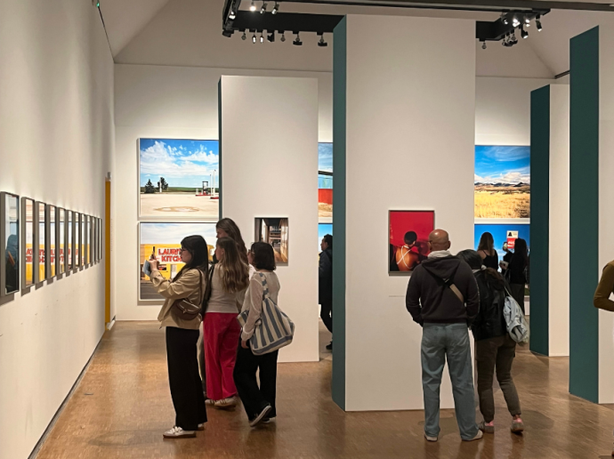 Des visiteurs parcourent une galerie d'art moderne, observant des photographies colorées accrochées aux murs blancs. Plusieurs œuvres représentent des paysages lumineux et des portraits. Le sol est en bois clair, et des spots au plafond éclairent les tableaux. Les visiteurs semblent attentifs et certains prennent des photos ou discutent devant les œuvres.