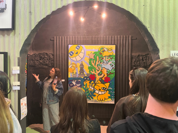 Une femme fait une présentation devant un groupe de personnes dans une galerie d'art. Elle se tient près d'une grande peinture colorée représentant des chats jaunes stylisés, la tour Eiffel et divers éléments floraux et abstraits. L'œuvre est accrochée sous un éclairage en arc, accentuant les couleurs vives. L'ambiance est conviviale et attentive.