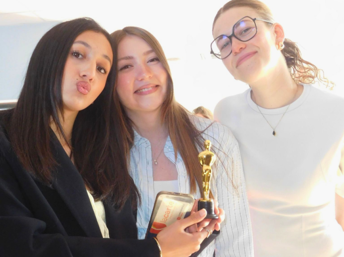 Trois jeunes femmes souriantes posent ensemble pour une photo. Celle à gauche fait une moue en direction de l'appareil, tandis que les deux autres sourient. La femme au centre tient un petit trophée doré en forme de statuette ainsi qu'une boîte. Elles semblent célébrer un moment festif ou une remise de prix.