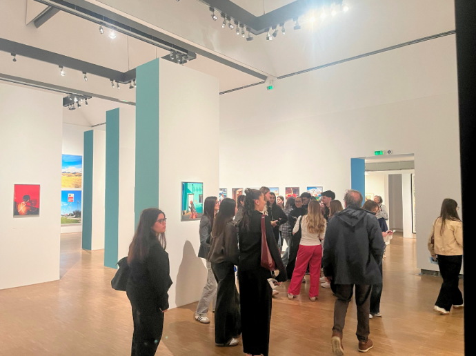Un groupe de personnes visite une exposition d'art moderne dans une galerie lumineuse aux murs blancs et colonnes vert pâle. Plusieurs œuvres colorées sont accrochées aux murs. Les visiteurs, de différents âges, discutent et observent les tableaux. Le sol est en bois et l'éclairage met en valeur les œuvres exposées.