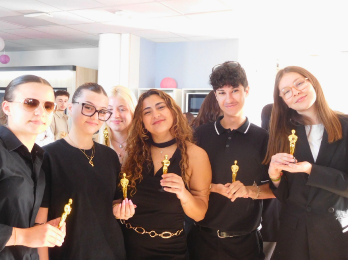 Six jeunes personnes debout côte à côte, souriantes, tenant chacune un petit trophée en forme de statuette dorée. Elles portent principalement des vêtements noirs et semblent être dans une pièce lumineuse et moderne, décorée de quelques ballons. L'ambiance est joyeuse et conviviale, comme lors d'une remise de prix ou d'un événement festif.
